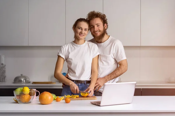 Una familia feliz en su nueva casa. Hombre guapo abrazando esposa sonriendo mirando a la cámara. Hermosa pareja joven hablando en videollamada usando computadora portátil. Pareja joven cocinando comida saludable en la cocina en casa — Foto de Stock