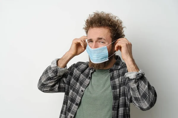 Disgustedly under duress puts on protective medical mask on his face to protect coronavirus, with curly hair, young man wearing plaid shirt and olive t-shirt under. White background — Fotografia de Stock