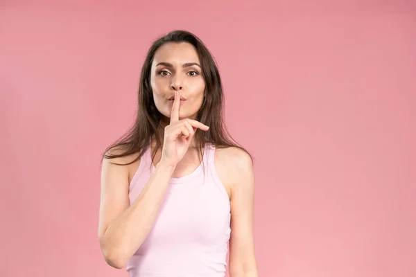 Gesture with a finger to the mouth says to be quiet or keep a secret young woman with dark hair in trendy pink t-shirt looking at camera. Facial expressions, emotions, feelings — Fotografia de Stock