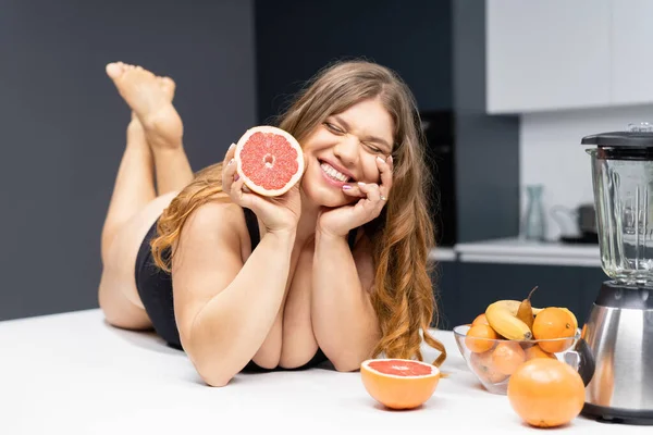 Young sexy chubby white girl in black swimsuit, white shirt at modern kitchen table. juggling fruits in hand. Trying to loose weight fast. Girl with obese problem. Fat barefoot girl low carb diet