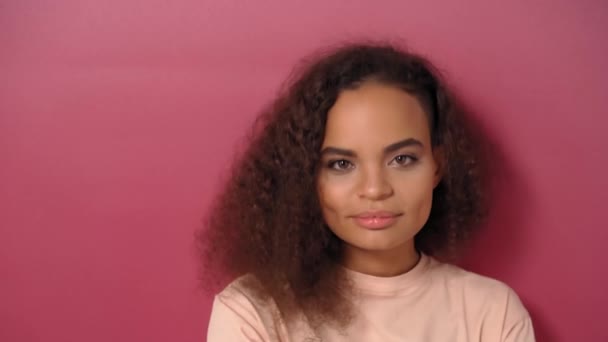 Urocza, uśmiechnięta kiwająca głową Afroamerykanka z długimi kręconymi włosami, patrząca pozytywnie w kamerę w wyluzowanej koszulce na różowym tle. Materiał 4K — Wideo stockowe