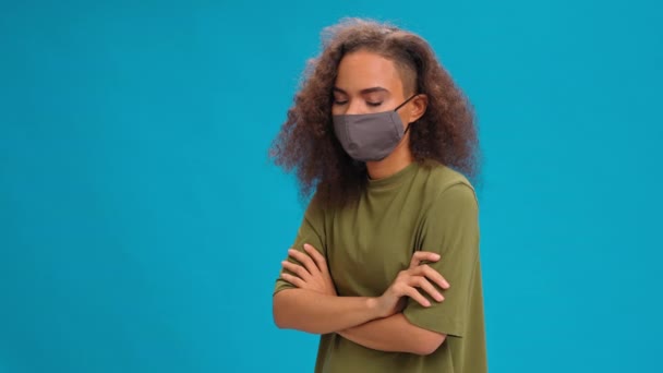 De pé com as mãos dobradas dizendo NÃO cabeça balançando menina muito afro-americana em máscara protetora reutilizável. Falando na câmera isolada no fundo azul — Vídeo de Stock