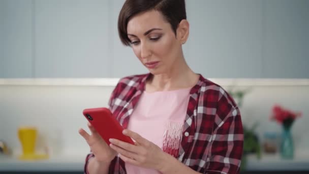 プレイしたシャツを着て夕食を調理しながら、モダンな明るいキッチンに立って彼女の赤いスマートフォンを使用してテキスト。健康的な食べ物を残す-ビーガンコンセプト.FHD映像 — ストック動画