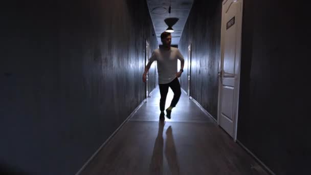 Νέος cool άνθρωπος σύγχρονο hip hop χορευτής εκτελεί το χορό του στην κάμερα. Χορεύοντας σε μια αχτίδα φωτός σε ένα μακρύ στενό σκοτεινό διάδρομο με πολλές λευκές πόρτες και στις δύο πλευρές. Βίντεο 4K — Αρχείο Βίντεο