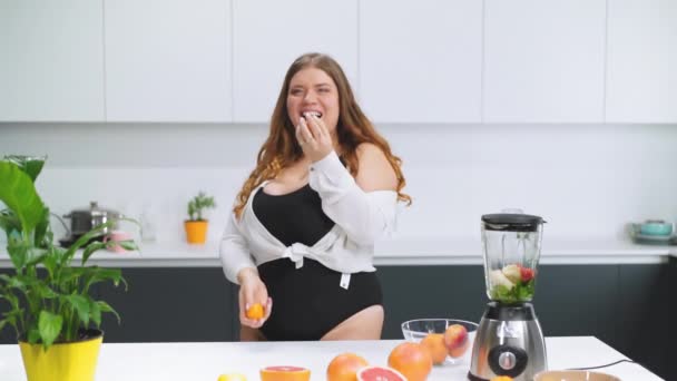 Happy tossing chaotically tangerines curvy body girl with long blond hair wearing black swimsuit making fresh fruits for healthy smoothie. Dieting and nutrition concept. File High Definition — Stock Video