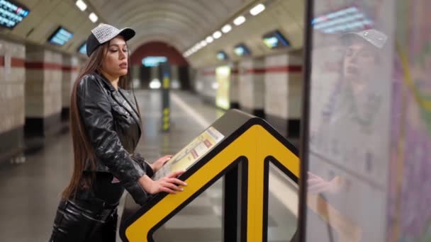 Sexy ragazza in giacca di pelle appoggiata su stand informativo guardando la fotocamera in metropolitana o metropolitana piattaforma al rallentatore. FHD. Metropolitana di Kiev, Kiev, Uraine. dicembre 2020 — Video Stock