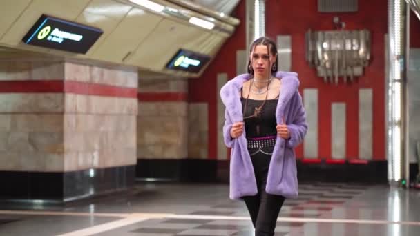 합성 자주색 모피 코트를 입은 패션젊은 여성이 지하철이나 지하철 승강장에서 천천히 걸을 때 매력적으로 걷는 모습을 어깨에 드러냈다. 화려 한 화장을 한 여성. FHD. Kyiv Metro, Kyiv, Uraine. 2020 년 12 월 — 비디오