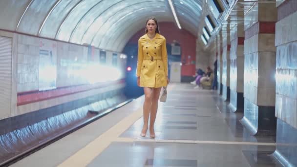 Berjalan seksi di peron dengan kereta yang akan datang, gadis muda cantik dengan kaki panjang dengan mantel musim semi kuning dan tas putih di tangannya. FHD. Kyiv Metro, Kyiv, Uraine. Desember, 2020 — Stok Video
