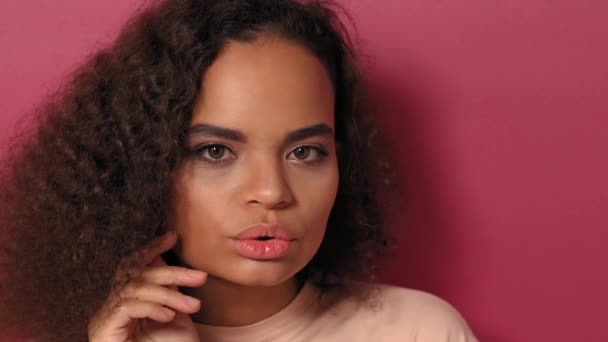 美しいアフリカ系アメリカ人の少女は、ピンクの背景に隔離された桃のシャツを着てカメラをそっと顔に触れている。美しさの概念。4K映像 — ストック動画