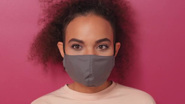 Speelse, koketterige, lastige look met herbruikbaar gezichtsmasker Afro-Amerikaans charmant meisje in perzikkleurig t-shirt, om te voorkomen dat van corona COVID-19 en SARS cov 2 infectie geïsoleerd op roze achtergrond Rechtenvrije Stockvideo's