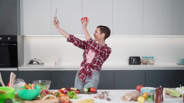 Mujer bonita tomando selfie usando teléfono inteligente sosteniendo pimiento morrón de pie en su nuevo hogar mientras cocina ensalada fresca con una camisa a cuadros con un estilo de pelo bob. Imágenes de FHD — Vídeo de stock