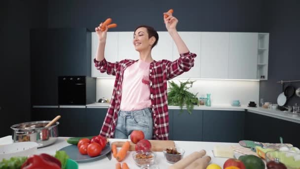 Наслаждаюсь домашней жизнью, танцуя с морковью в руках женщины в новой кухне нового дома. Счастливая женщина танцует с морковью в руках. Счастливая женщина наслаждается приготовлением еды. Кадры FHD — стоковое видео