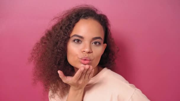 Küsse an andere junge Afroamerikanerin mit lockigem Haar, die positiv in die Kamera blickt und ein pfirsichfarbenes T-Shirt auf rosa Hintergrund trägt. Beauty-Konzept. 4K-Filmmaterial — Stockvideo