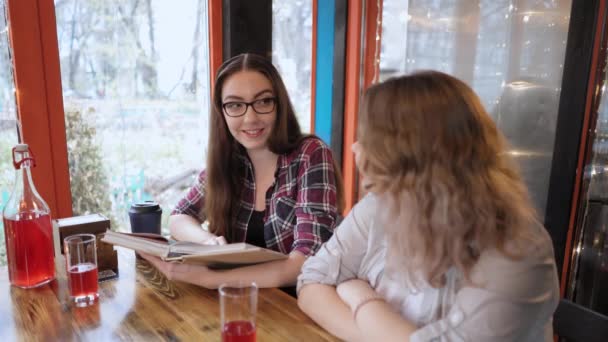 Kahve molası sırasında kafede sohbet eden başarılı çekici bayan arkadaşlar arka planda kitap okuyan ışıklar eşliğinde çalışma notlarını paylaşıyorlar. 4K görüntü — Stok video