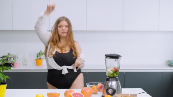 食事と栄養の概念。黒のレオタードと白いシャツに身を包んだ幸せな太りすぎの女の子を踊る。新鮮なフルーツジュースを準備キッチンカービーボディガールブロンディで踊る。FHD映像 — ストック動画