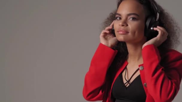 Feliz chica afroamericana joven escuchando música en auriculares con chaqueta roja top negro bajo aislado en fondo gris, emocionalmente se mueven, se divierten. Imágenes 4K — Vídeos de Stock