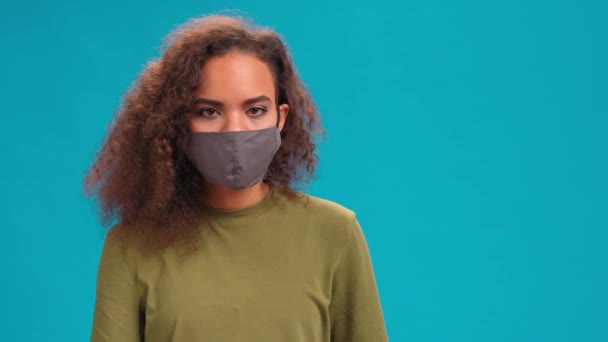 Testa annuendo indossando maschera viso riutilizzabile afro-americana affascinante ragazza in t-shirt di ulivo. Prevenire da COVID-19 corona e SARS cov 2 infezione isolata su sfondo blu. Filmati 4K — Video Stock