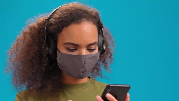 Afro-Amerikaans meisje met een smartphone draagt een medisch gezichtsmasker om de verspreiding van het coronavirus te voorkomen. Meisje in olijf t-shirt geïsoleerd op blauwe achtergrond. 4K-beelden Stockvideo