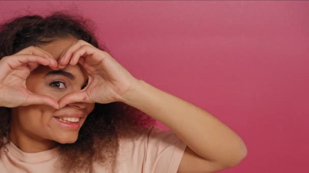 Patrząc przez miłość lub gest serca piękna młoda Afroamerykanka uśmiecha się pozytywnie do kamery nosząc brzoskwiniowy t-shirt odizolowany na różowym tle. Koncepcja miłości. Nagranie 4k — Wideo stockowe
