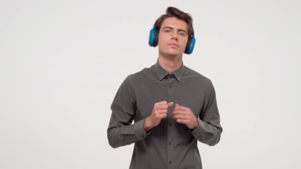 Nieśmiały tancerz młody człowiek słuchający muzyki w niebieskich bezprzewodowych słuchawkach ubrany w szary strój sportowy. Zabawny, tańczący młodzieniec odizolowany na białym tle. Materiał 4K — Wideo stockowe