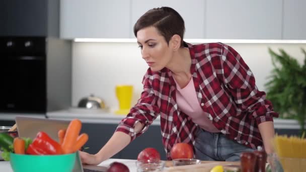 Jovem mulher usando laptop na cozinha procurando receitas on-line ou assistindo show on-line enquanto cozinha. Mulher jovem cozinhar na cozinha de uma nova casa. Vida saudável a viver. câmara lenta. Imagens de FHD — Vídeo de Stock