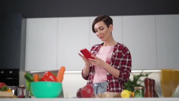 中年の女性は、健康的な食品を調理し、オンラインレシピを見て.台所の主婦が携帯電話をタイプしている。主婦の概念。高品質 — ストック動画