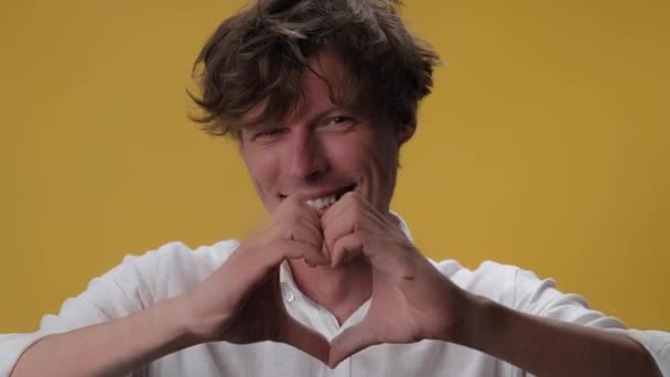 Young man making heart shaped hands gesture on isolated on yellow background. The winking face of a handsome young man in a white shirt. High quality 4k video — Stock Video