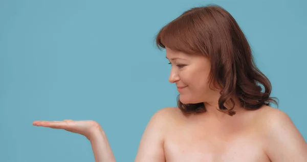 Veselá zralá žena držící otevřenou ruku pro vizualizaci péče o pleť. Usmívající se žena inzerovat anti-age krém izolované na barevném pozadí — Stock fotografie