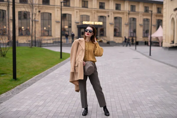 Charmante fille modèle dans un manteau beige d'automne et des lunettes de soleil, chapeau panama à carreaux debout avec le visage tourné tenant manteau sur ses épaules. Photo tonique rétro — Photo