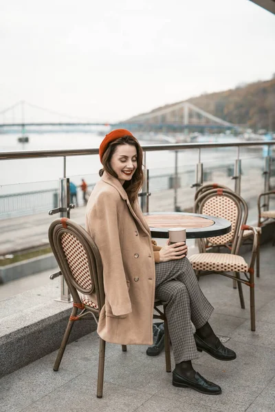 Preciosa joven francesa sentada en la terraza del restaurante con taza de café mirando a la cámara. Retrato de una mujer joven y elegante con abrigo de otoño y boina roja al aire libre —  Fotos de Stock