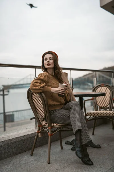 Preciosa joven francesa sentada en la terraza del restaurante con taza de café mirando a la cámara. Retrato de una mujer joven y elegante con abrigo de otoño y boina roja al aire libre —  Fotos de Stock
