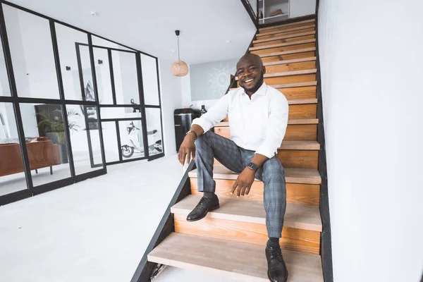 Glad ung framgångsrik affärsman i sitt köpta hus. Afro American Sits På Trappor I Hall i lägenheter och leenden. Begreppet personlig tillväxt, framgång, övervinna svårigheter — Stockfoto