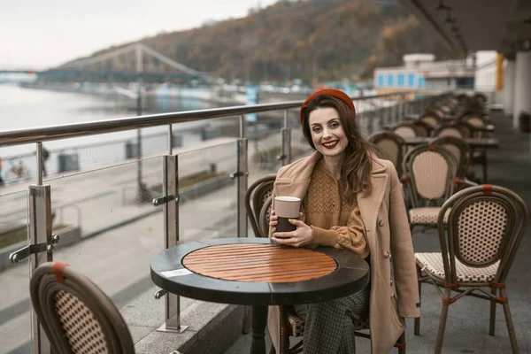 Açık hava lokantasında ya da terasta oturan mutlu genç kız elinde kahve fincanıyla sırıtıyor. Sonbahar ceketi ve açık havada kırmızı bereli şık bir genç kadının portresi. — Stok fotoğraf