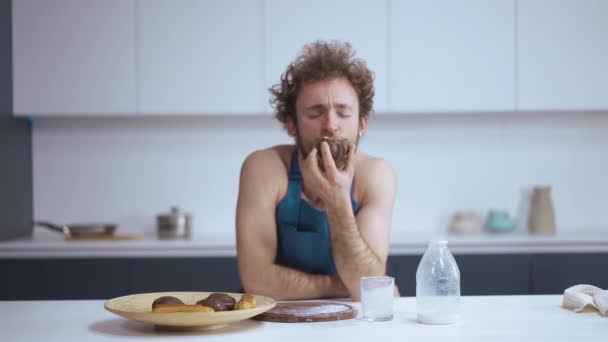 Young sexy guy eating a cupcake. A curly haired young man in a blue denim apron is having breakfast. Sitting in kitchen at kitchen table — Wideo stockowe