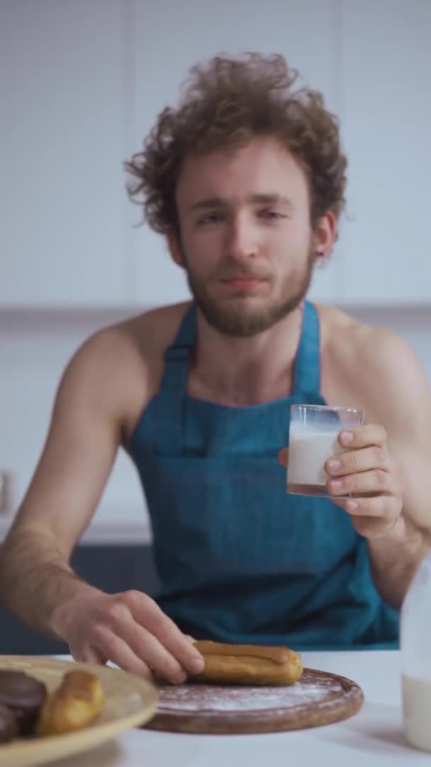 Młody, seksowny Francuz je eklerkę i pije mleko ze szklanki w kuchni. Kręcony młody człowiek w dżinsowym niebieskim fartuchu je śniadanie w porannym świetle — Wideo stockowe