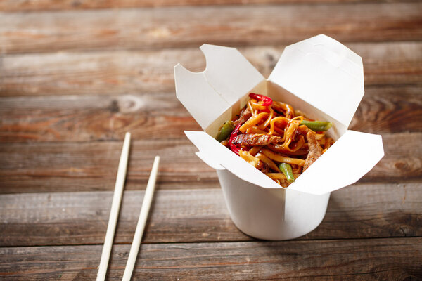 Noodles with pork and vegetables in take-out box on wooden table