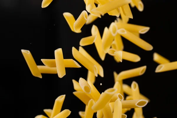 Fallende Penne-Pasta. Fliegende gelbe rohe Makkaroni über schwarzem Hintergrund. — Stockfoto