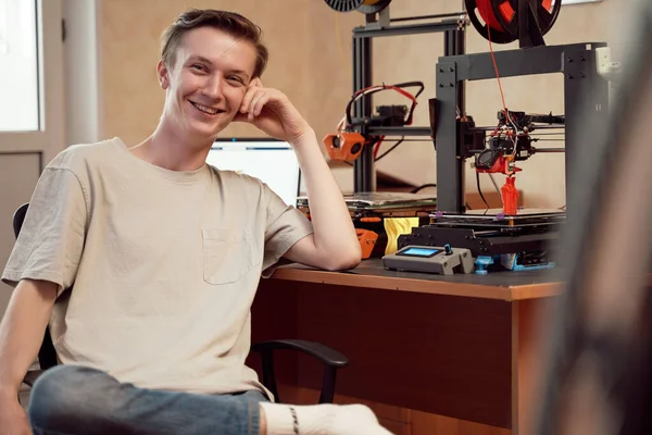 Веселый молодой человек сидит рядом с 3D принтерами и ноутбуком — стоковое фото