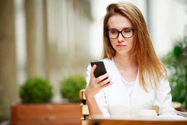 Chica mensajes de texto en el teléfono inteligente en una terraza del restaurante con un fondo desenfocado —  Fotos de Stock