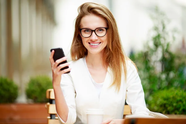 Chica mensajes de texto en el teléfono inteligente en una terraza del restaurante con un fondo desenfocado —  Fotos de Stock