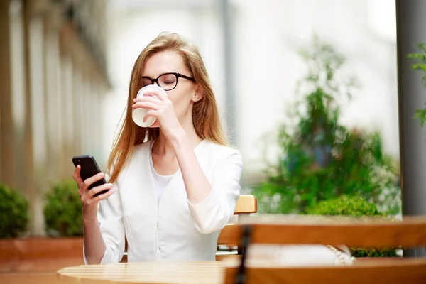 Chica mensajes de texto en el teléfono inteligente en una terraza del restaurante con un fondo desenfocado —  Fotos de Stock