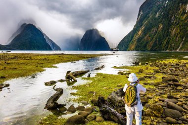  Sırt çantalı, resimli manzaralı yaşlı bir kadın. Milford Sound fiyort kıyısı bataklık çimleriyle kaplandı. Yeni Zelanda. Egzotik, aktif ve fotografik turizm kavramı