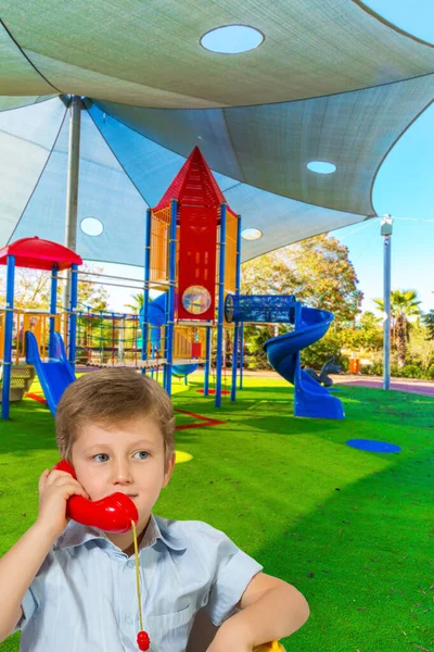 Charming blond boy talking on the red phone. Cosy children \'s playground with a variety of multicolored attractions. The concept of physical and mental development of children