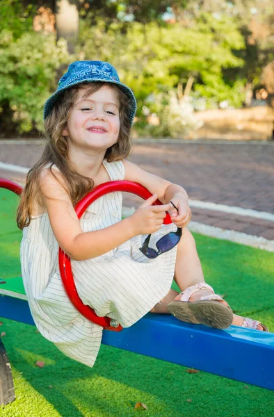 迷人的小女孩穿着巴拿马式的长发 在孩子们的秋千上欢快地笑着 温暖的夏日 在操场上 — 图库照片
