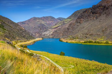 Tepelerin ve çimenlerin arasındaki gölün gök mavisi derin suları. Cromwell kasabası yakınlarındaki Dunstan Gölü. Güney Adası, Yeni Zelanda. Ekolojik ve fotoğraf turizmi kavramı