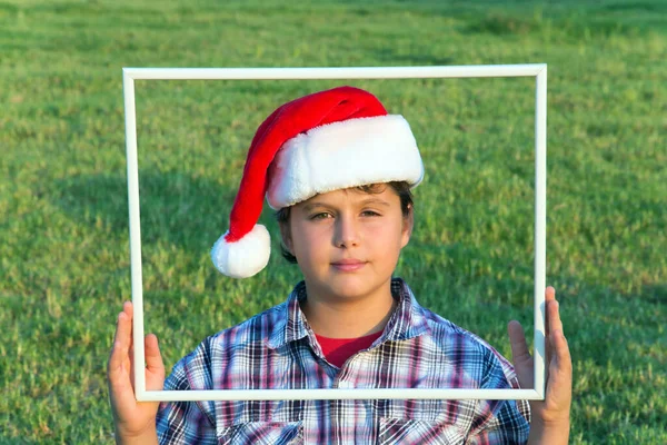 头戴小丑式圣诞帽的男孩正透过白色的框架看过去 背景是一片绿色的草坪 概念肖像和广告照片 — 图库照片
