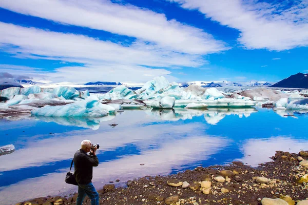 ジーンズを着た白髪の男が壮大な風景を撮影します アイスランド 白と青の氷山 氷の流れと雲はラグーンJokulsaurlounに反映されます 写真観光の概念 — ストック写真