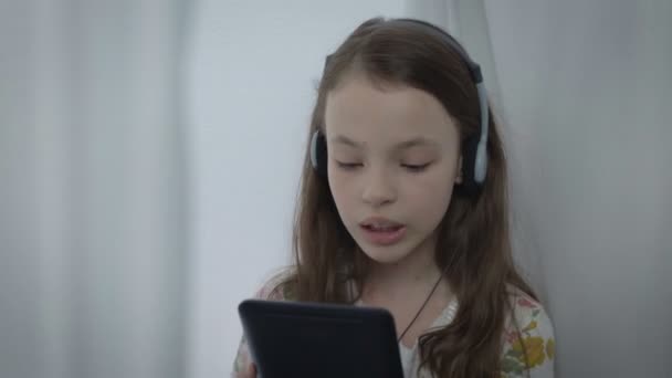 Śliczną dziewczynkę z słuchawki oglądając teledysk na tabletki i śpiewa. — Wideo stockowe