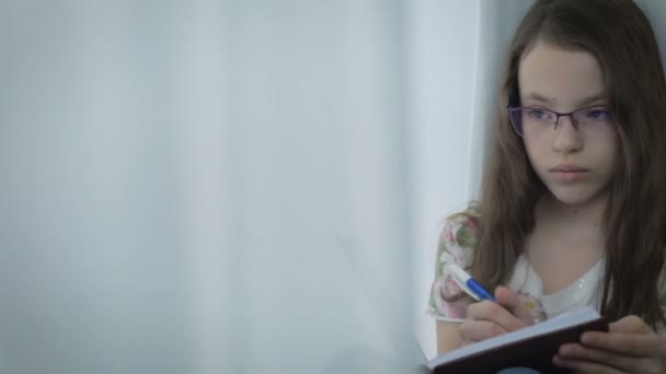 Niña seria en gafas diligentemente hace su tarea en la ventana . — Vídeo de stock