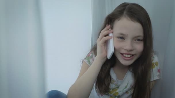 Schöne glückliche kleine Mädchen emotionale Gespräche auf dem Smartphone am Fenster. — Stockvideo